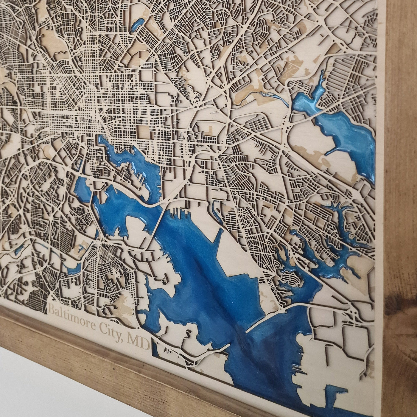 A framed laser-cut wooden city map wall art piece, showcasing detailed street etching with blue water features and a natural wood frame.