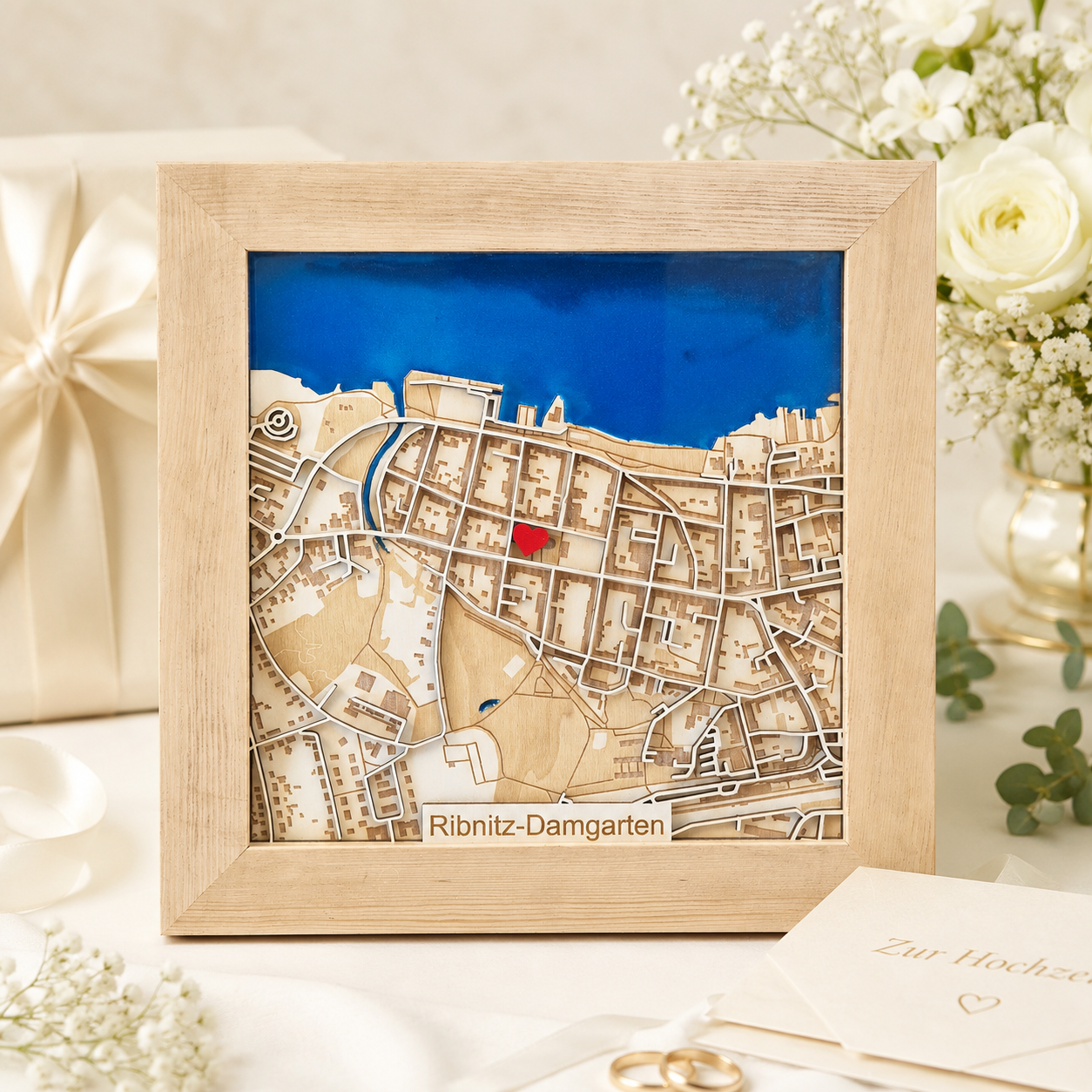 A framed wooden city map art piece of Ribnitz-Damgarten, featuring a light natural wood frame, beige and tan cutout streets and terrain, a bright blue background band, and a small red highlighted area. The framed piece is displayed on a white table with wedding-themed props including a gift box, flowers, and a card.