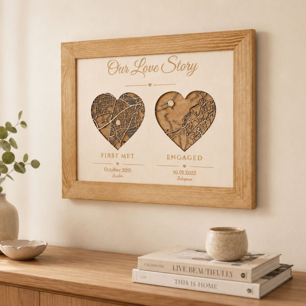 A framed personalized wooden map print titled 'Our Love Story' hangs on a cream wall above a wooden console table. The print features two heart-shaped map cutouts with engraved details, one labeled 'FIRST MET' and the other 'ENGAGED'.