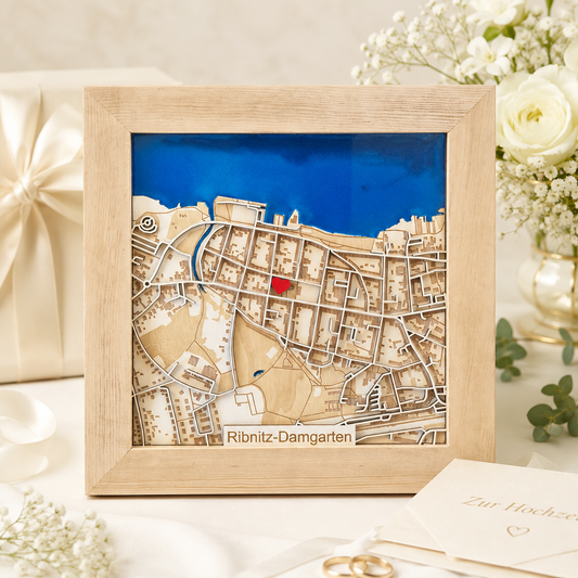 A framed wooden city map art piece of Ribnitz-Damgarten, featuring a light natural wood frame, beige and tan cutout streets and terrain, a bright blue background band, and a small red highlighted area. The framed piece is displayed on a white table with wedding-themed props including a gift box, flowers, and a card.