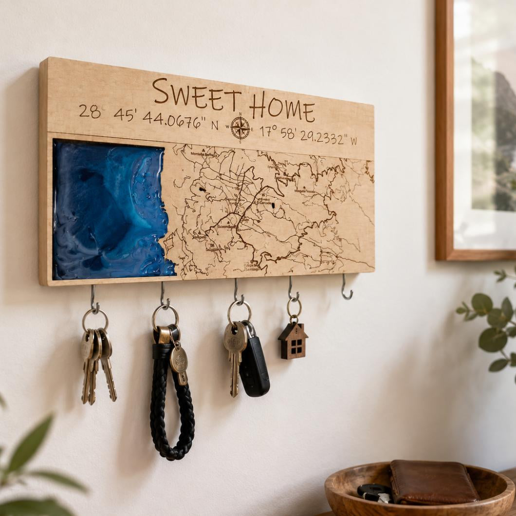 A personalized wooden wall-mounted key holder with engraved coordinates and the words “SWEET HOME,” featuring a map design on one side and a blue resin panel on the other. Five metal hooks below hold keys and a small house-shaped keychain, shown in a styled interior setting.