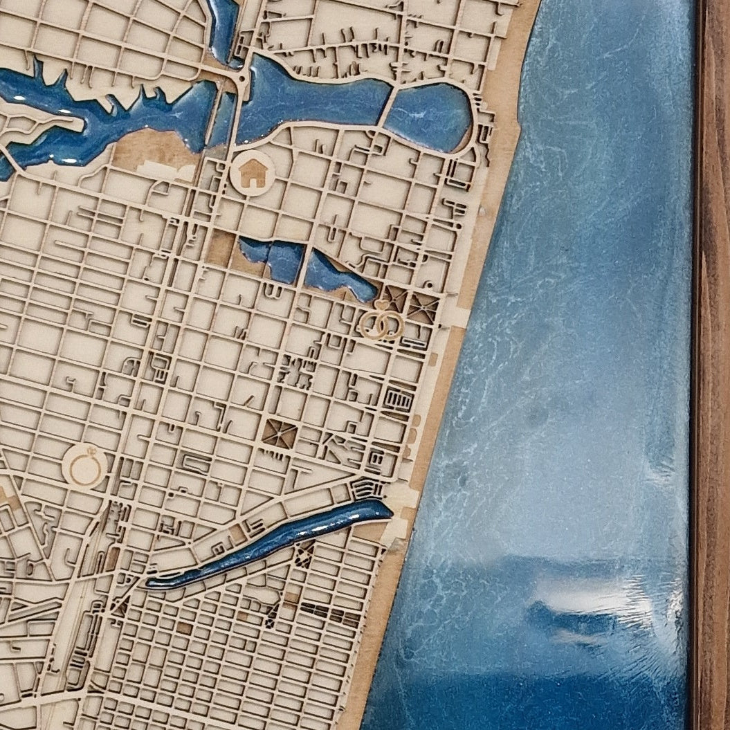 A close-up of a wooden city map wall plaque showing streets, blocks, waterways, and a large blue body of water along the right side, mounted against a wood surface.