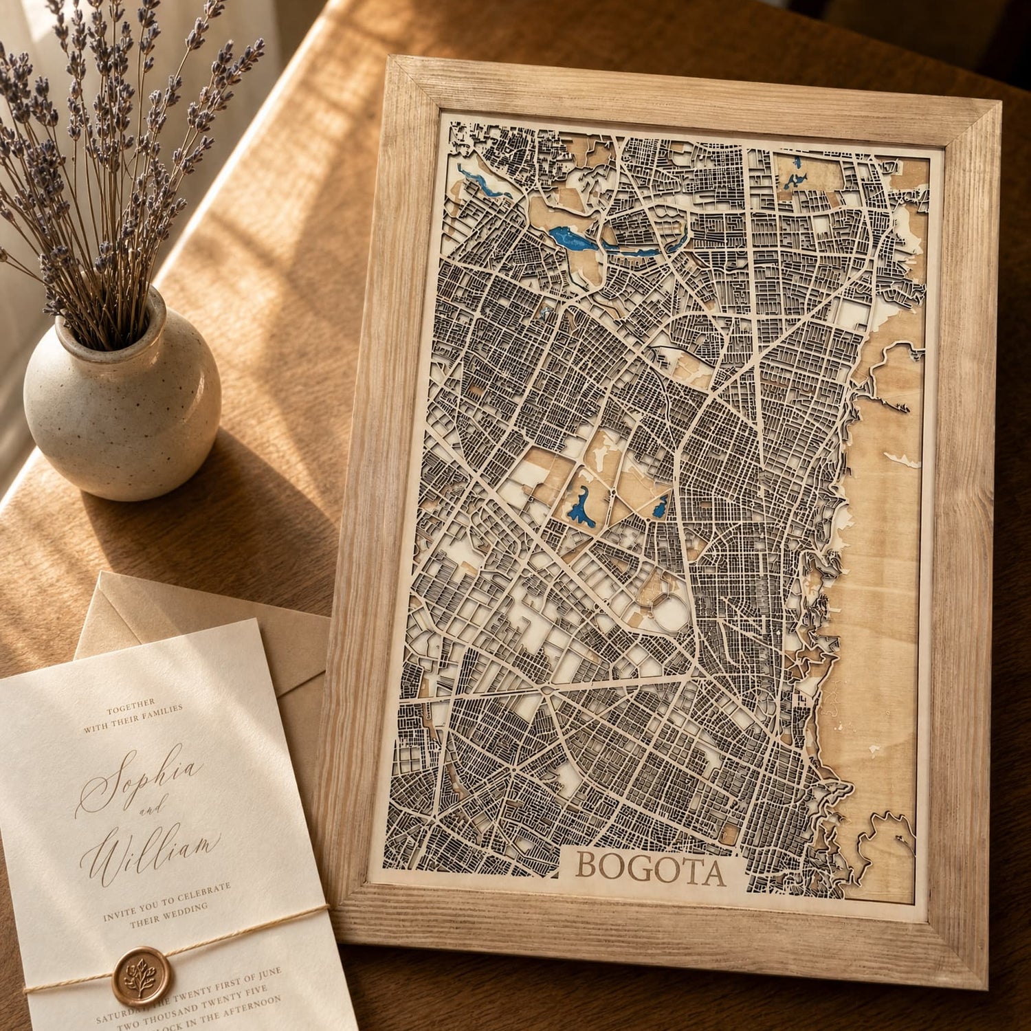 A framed wooden map print of Bogota, Colombia, styled on a warm wooden table next to a small vase of dried lavender and a wedding invitation card.