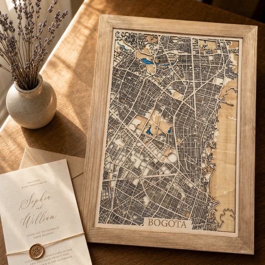 A framed wooden map print of Bogota, Colombia, styled on a warm wooden table next to a small vase of dried lavender and a wedding invitation card.