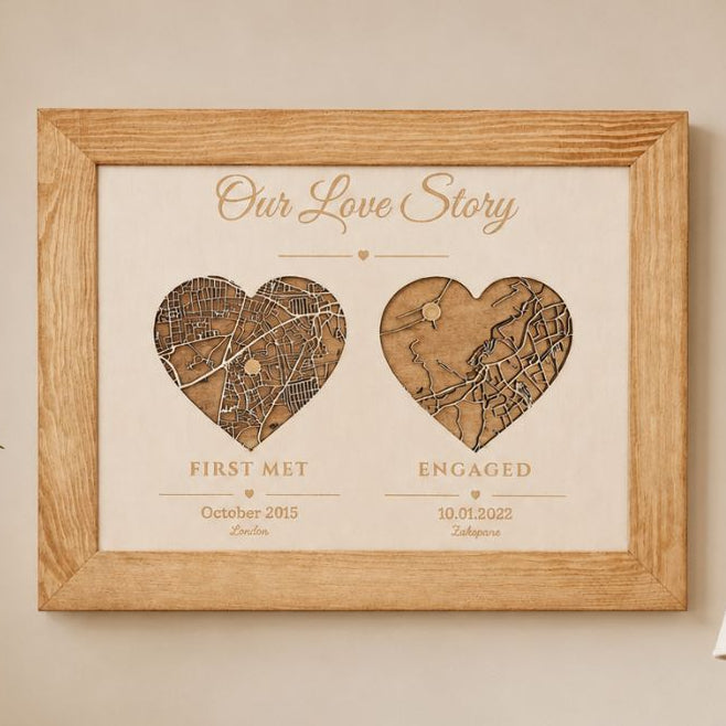 A framed personalized map art print with two heart-shaped map cutouts and the text 'Our Love Story' and 'First Meet' and 'Engaged' displayed on a wall above a wooden console table with a vase, books, and a lamp.